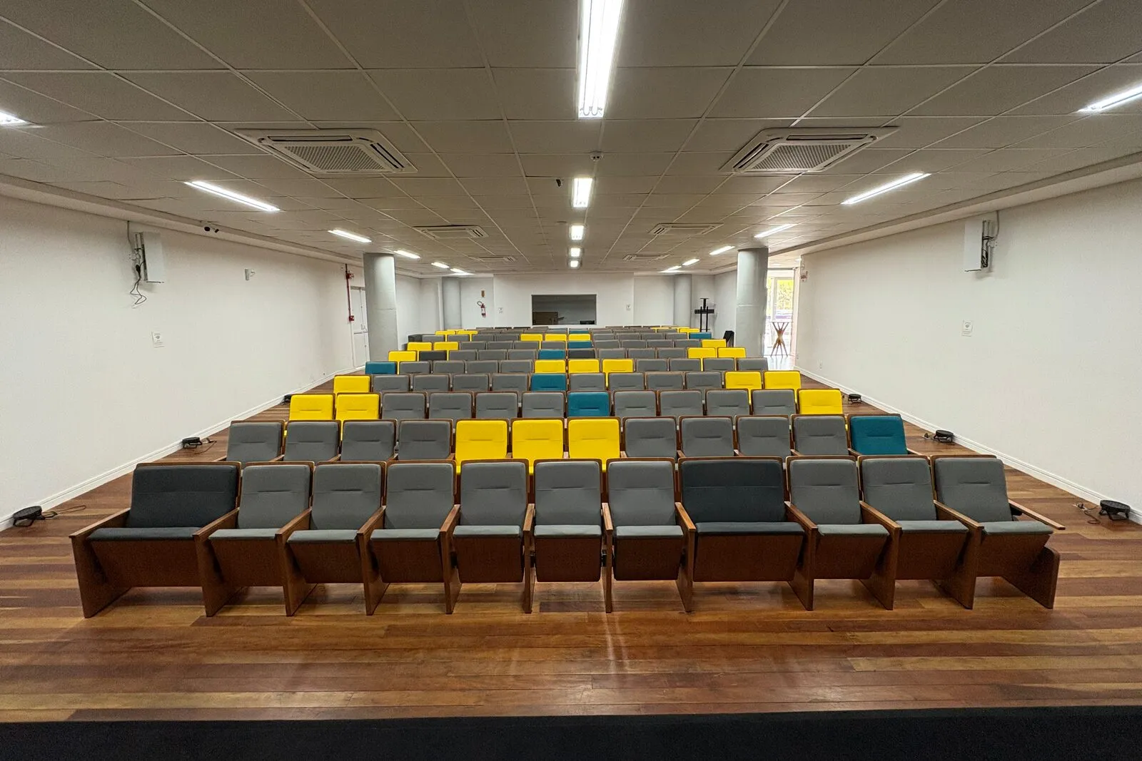 Auditório do Elume vazio, com poltronas cinza, amarelas e azuis em fileiras e piso de madeira