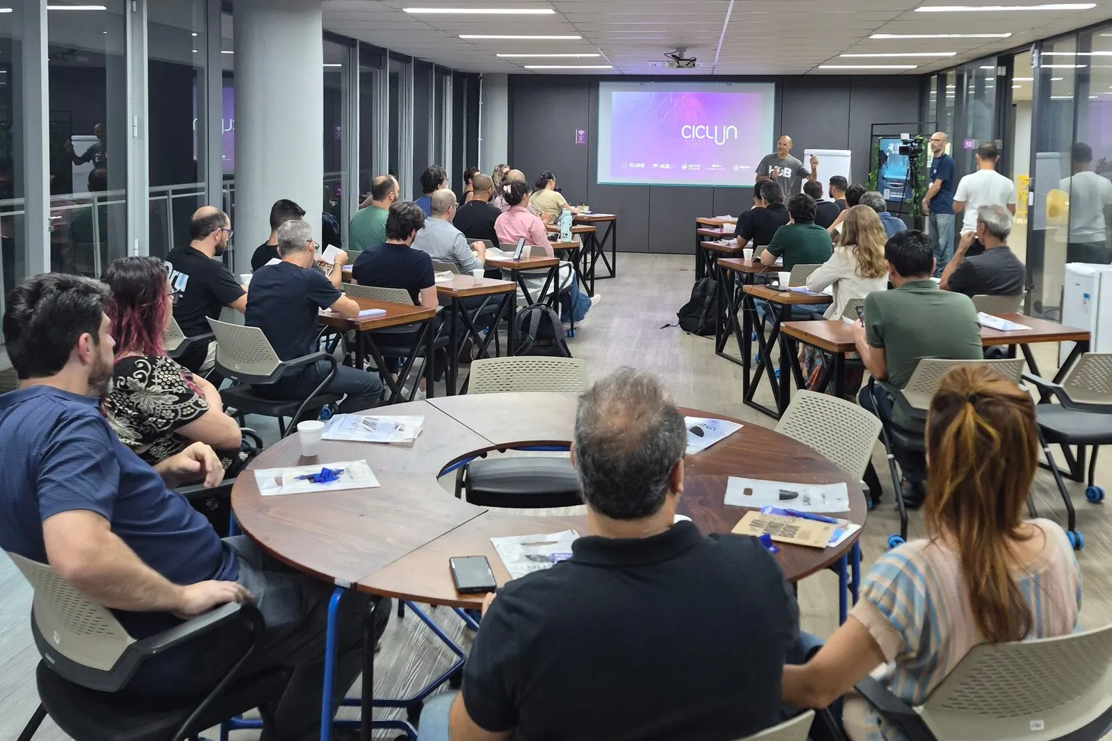 Plateia sentada em mesas redondas acompanha uma palestra no Elume, com apresentação projetada ao fundo