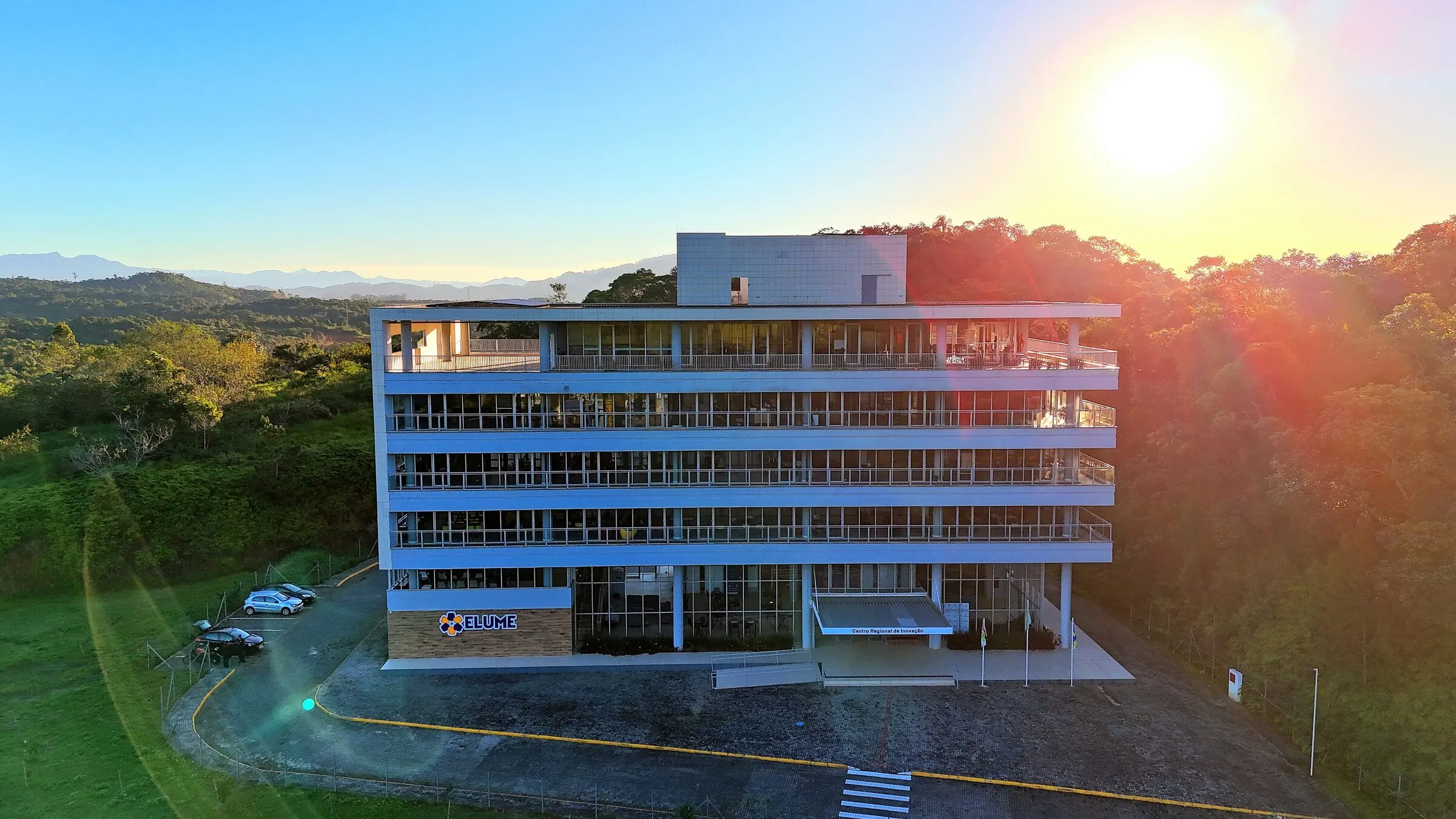 Vista aérea da fachada da sede do Elume Park ao pôr do sol, cercada por mata, em Itajaí