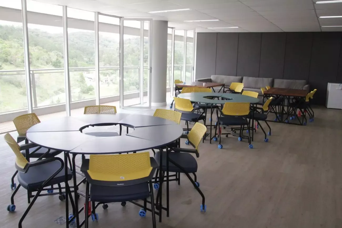 Sala multiuso do Elume com mesas redondas, cadeiras amarelas e vista para a mata